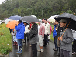 雨の中傘をさしながら説明を聞く参加者たち